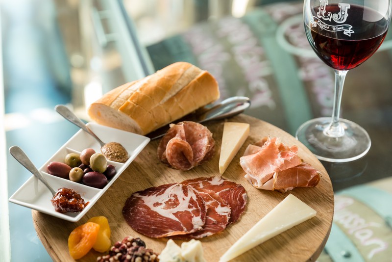 Platter of cheese, olives, bread, and cold meats beside a half filled glass of red wine.