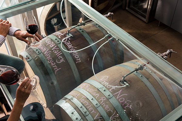 Two people sampling red wine from two barrels of wine.