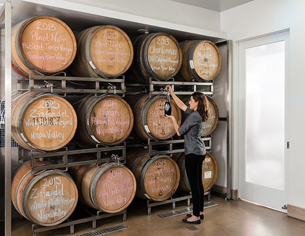A lady holding a growler of wine in front of 8 barrels of wine.