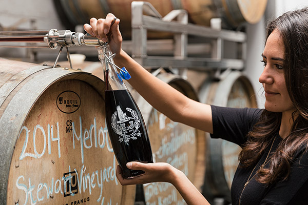 A lady filling red wine from a barrel into a refillable, eco-friendly growler. The growler has the La Jolla Crafted Wine logo on it.