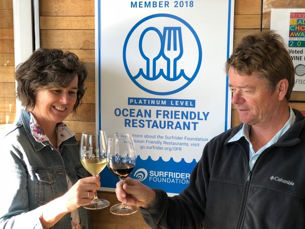 Lowell and Anne Jooste standing in front of a sign indicating "Platinum Level Ocean Friendly Restaurant"   recognition from Surfrider Foundation USA.