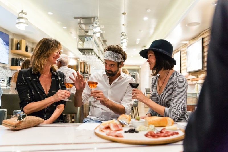 Three people enjoying wine from wine glasses.  A platter of bread, cheese and cold meats in   foreground.